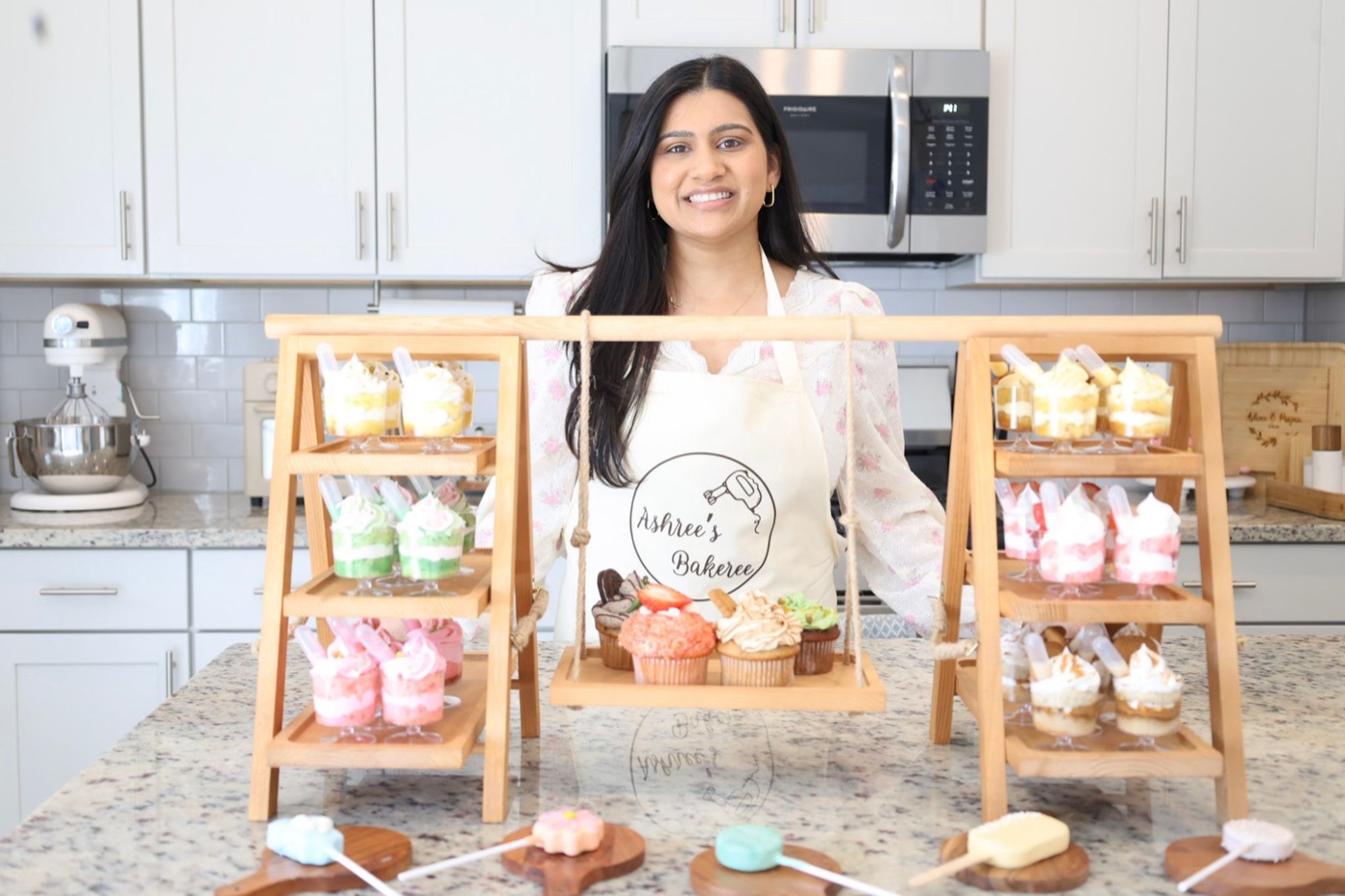Ashree, owner of Ashree's Bakeree, smiling behind a display of dessert cups, cupcakes, and cake pops in her kitchen