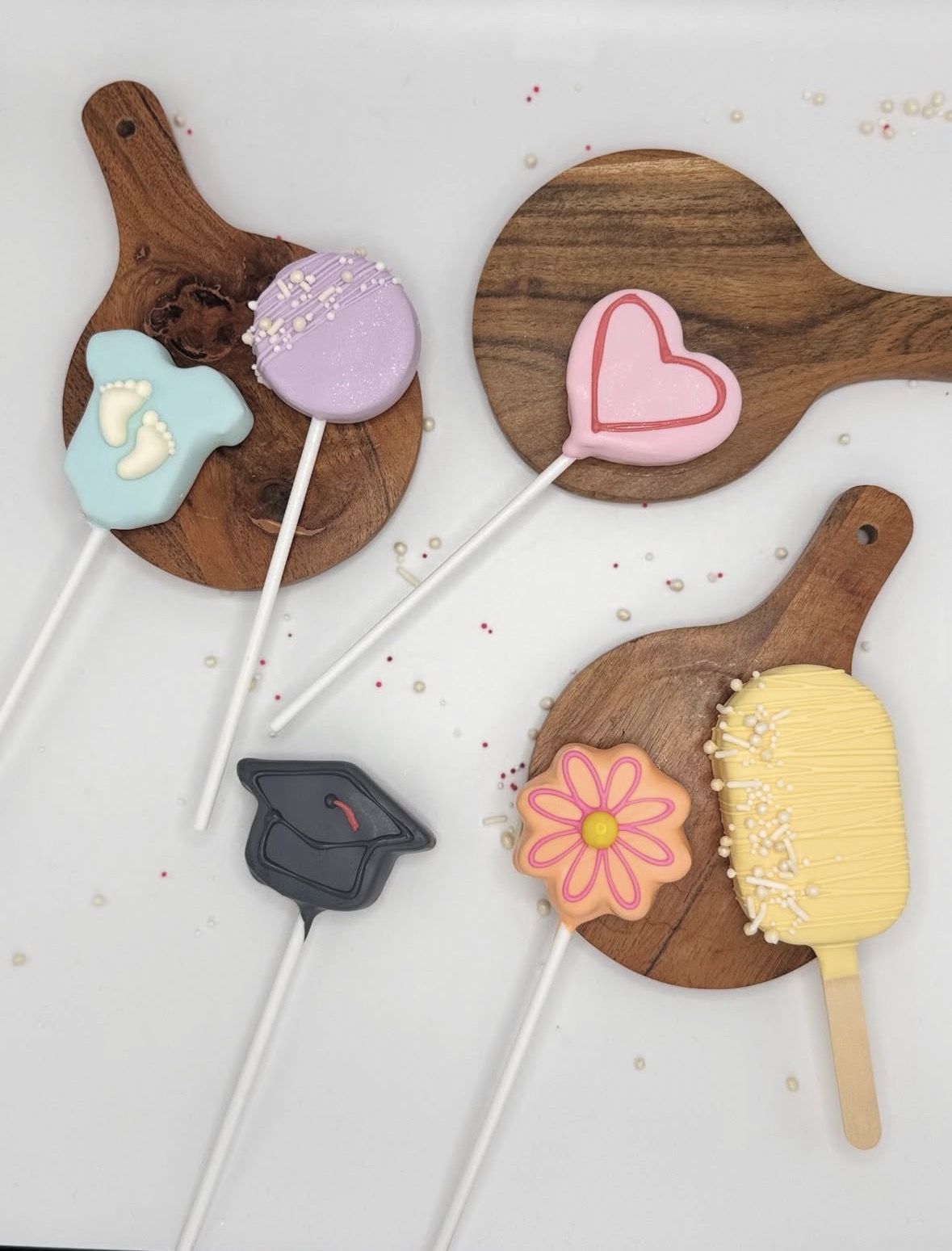 Assorted cake pops shaped as a baby onesie, heart, graduation cap, flower, and popsicle, each on a wooden board