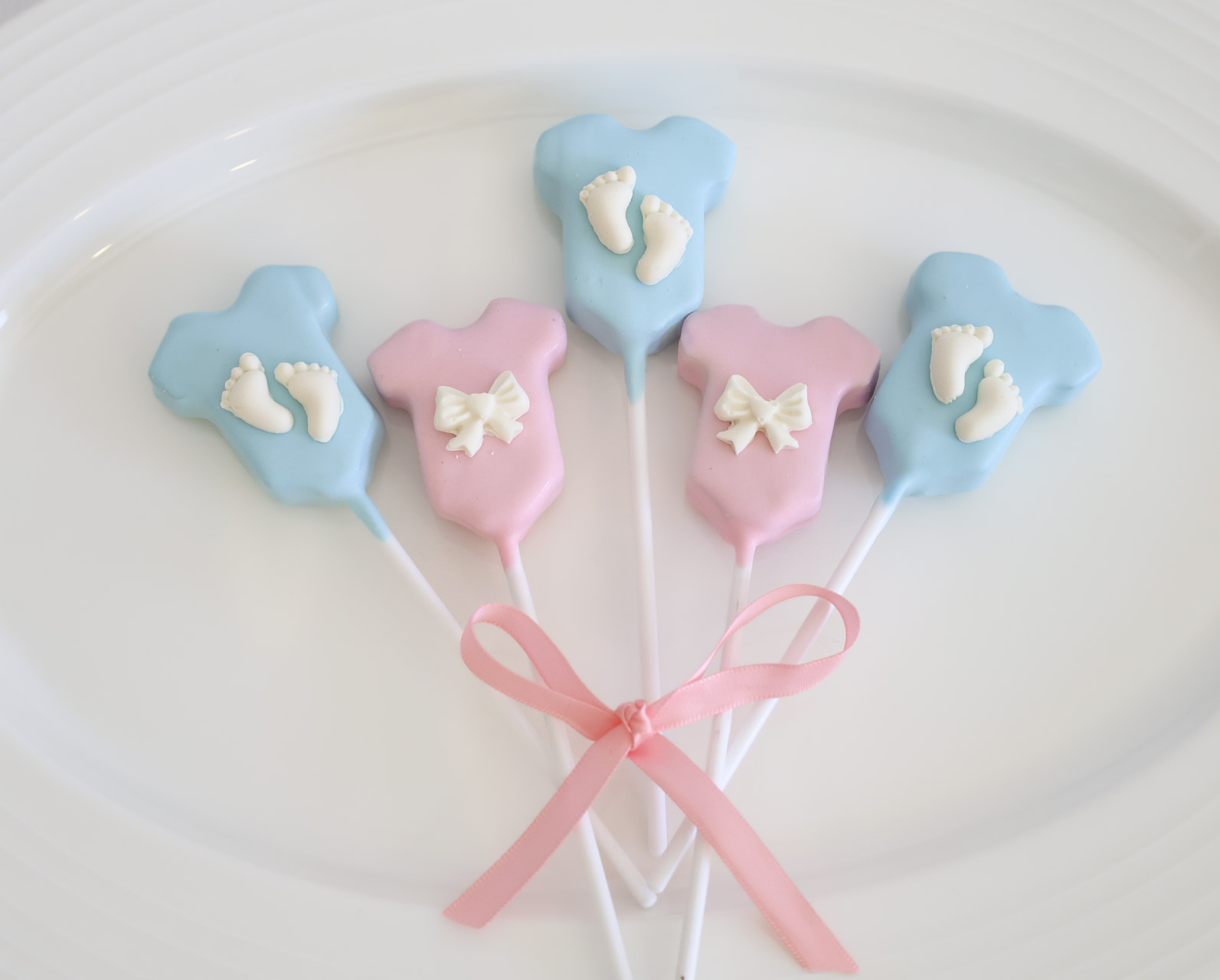 Blue and pink baby onesie shaped cake pops with tiny feet and bow details, tied with a pink ribbon