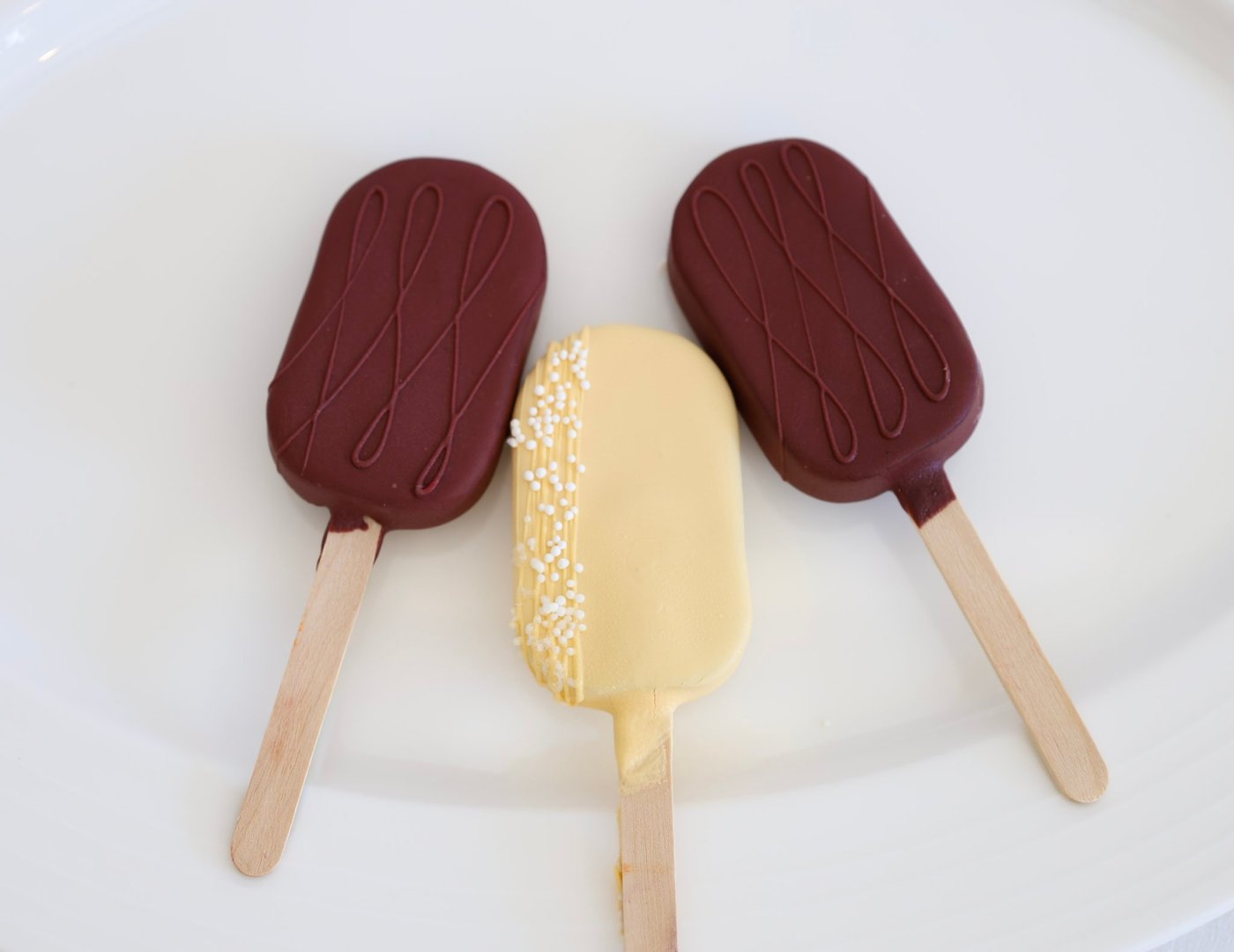 Three chocolate-dipped cakesicles, two in maroon and one in pale yellow with a nonpareil sprinkle stripe
