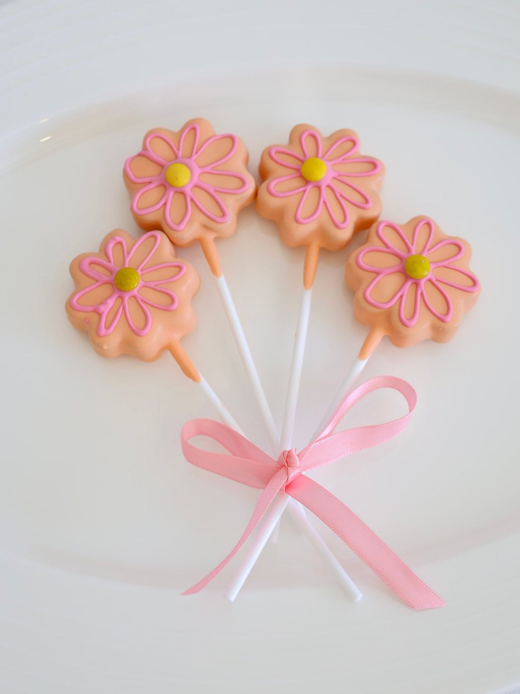 Four flower-shaped cake pops with pink daisy designs, tied together with a pink ribbon