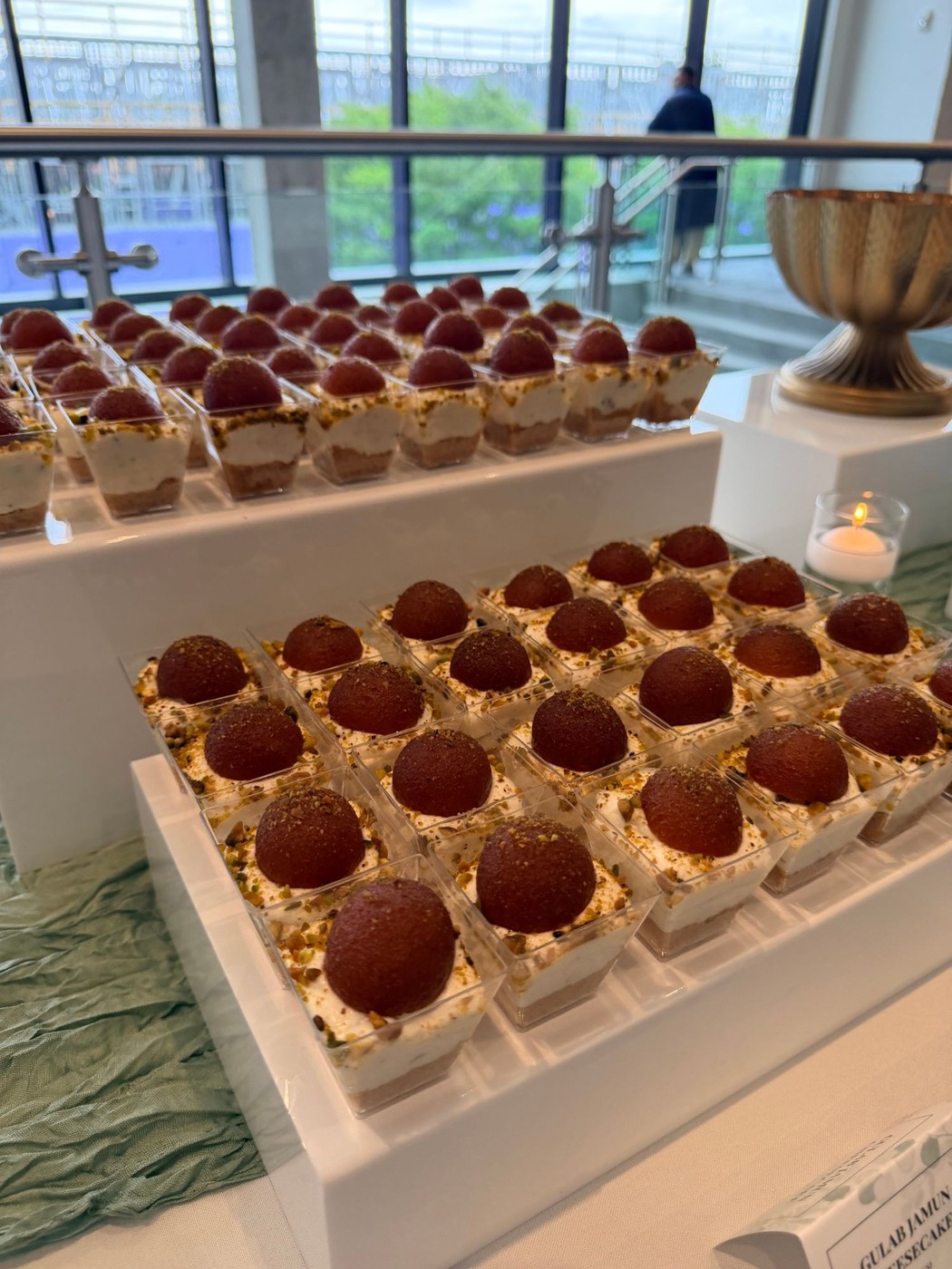 Rows of Gulab Jamun cheesecake shooters topped with pistachio, set up for a catering display