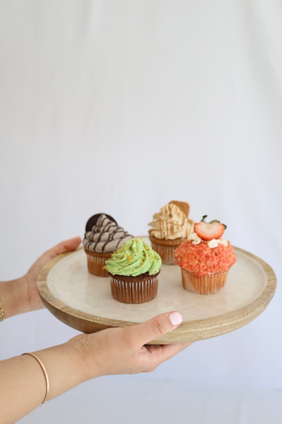 Cookies and cream, pistachio, Biscoff, and strawberry crunch cupcakes held on a round wooden board
