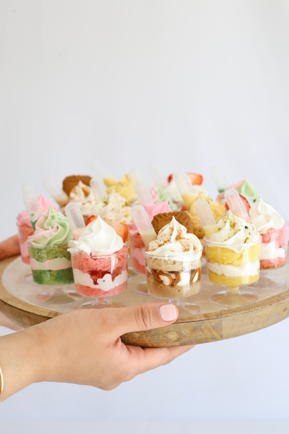 A hand presenting a wooden tray of assorted dessert cups; strawberry shortcake, pistachio, Biscoff, and lemon flavors, each topped with a dessert syringe