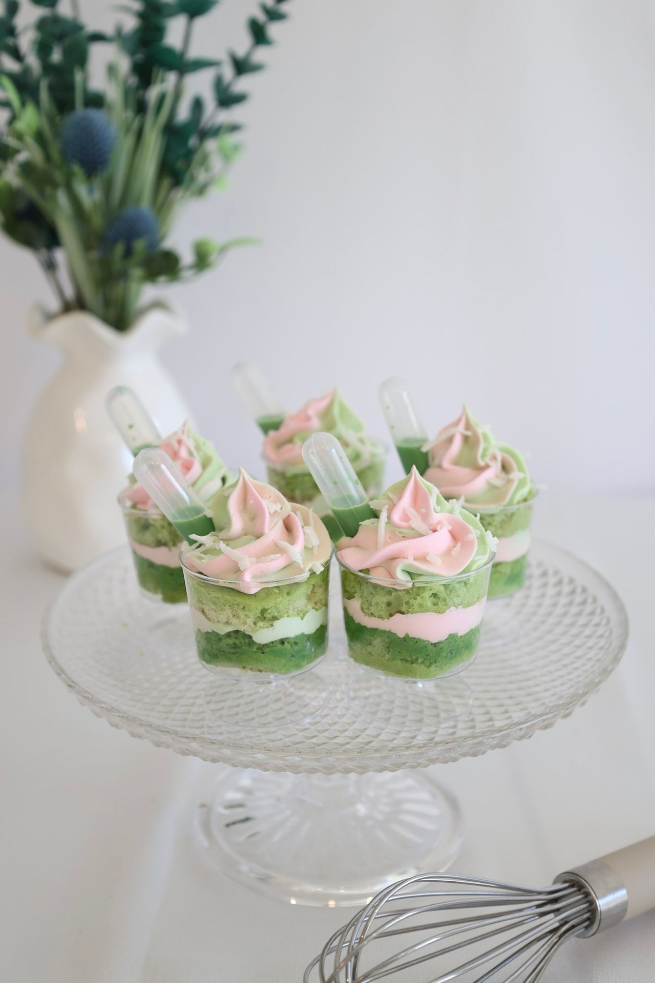 Matcha dessert cups topped with pink rose whipped cream and coconut flakes on a glass cake stand